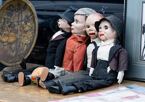 A trio of vintage ventriloquist dummies, each with distinct outfits and expressive faces, lined up at a flea market, waiting for their next act.