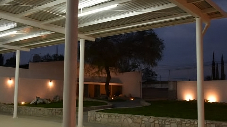 Vista nocturna del salón techado y el jardín de Villa Lanark, iluminados con luces cálidas amarillas en las jardineras y paredes.