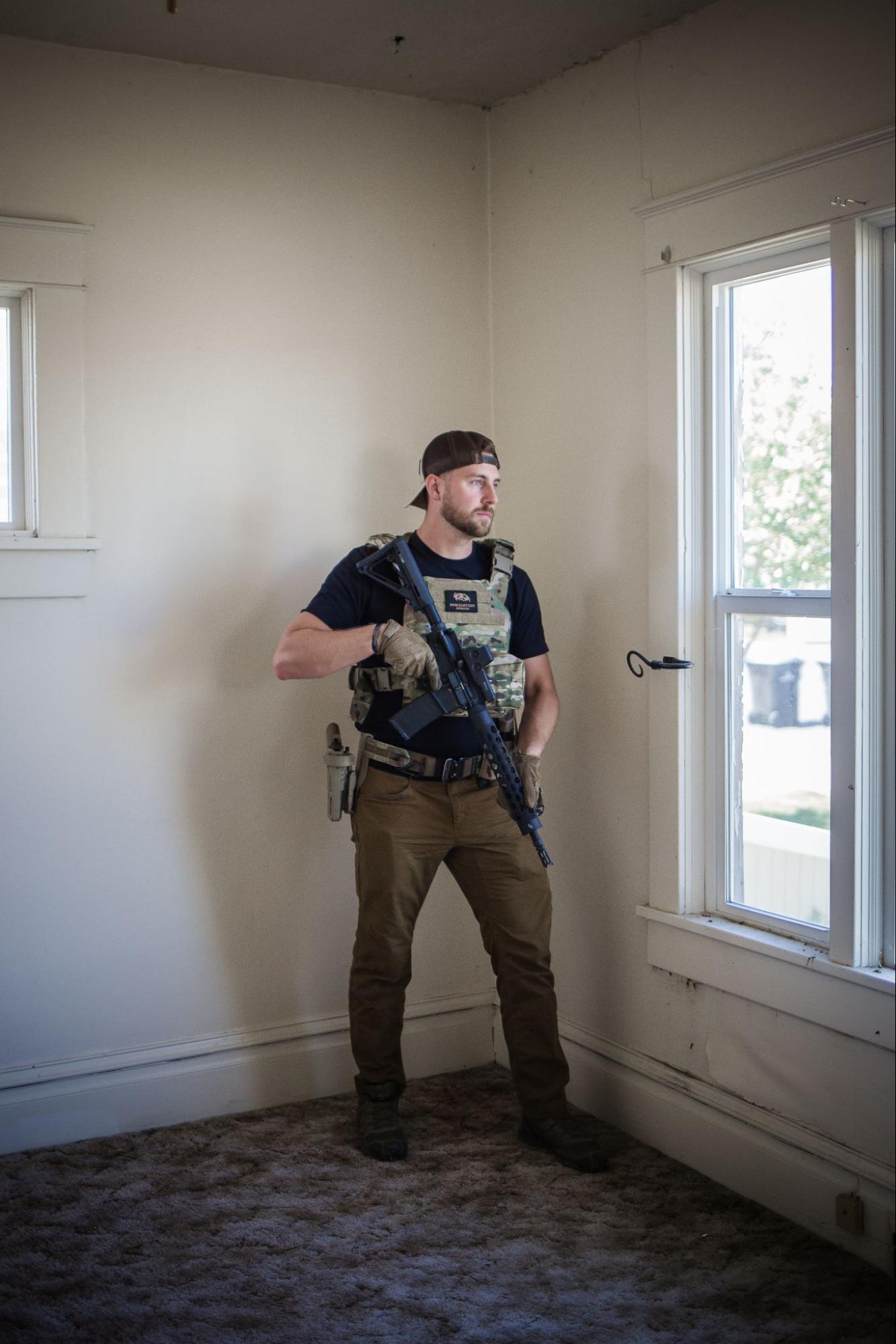 Armed man in tactical gear standing by a window in a plain room, looking outside
