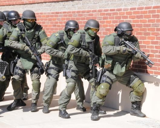 SWAT team in green tactical gear and helmets, holding rifles, advances along red brick wall, focused and alert in daylight.