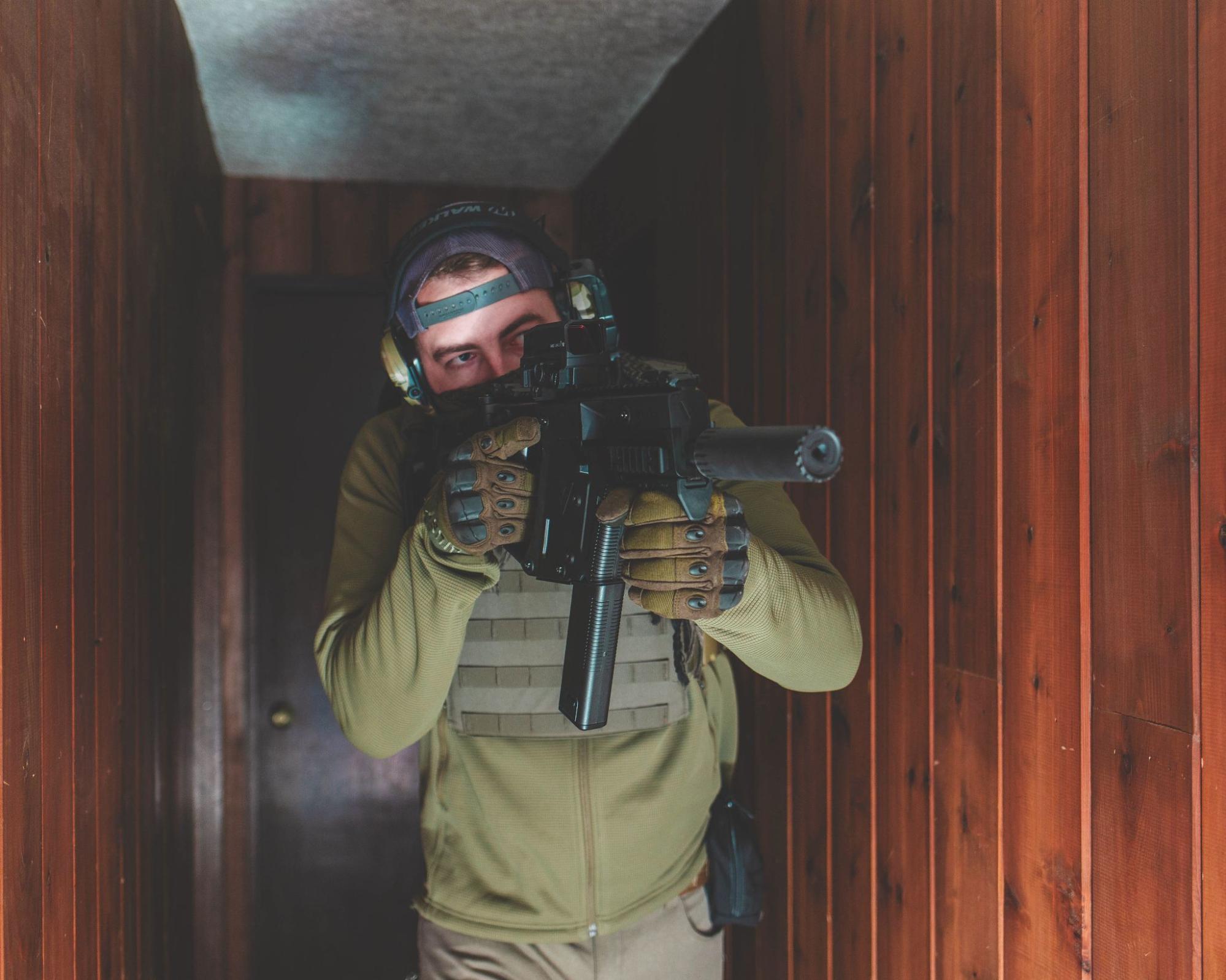 Person in tactical gear aims a rifle in a narrow wood-paneled hallway. Wearing ear protection and gloves, they appear focused and alert.
