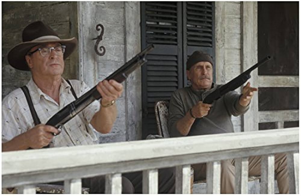 Two older men holding shotguns sit alertly on a rustic porch with wooden shutters