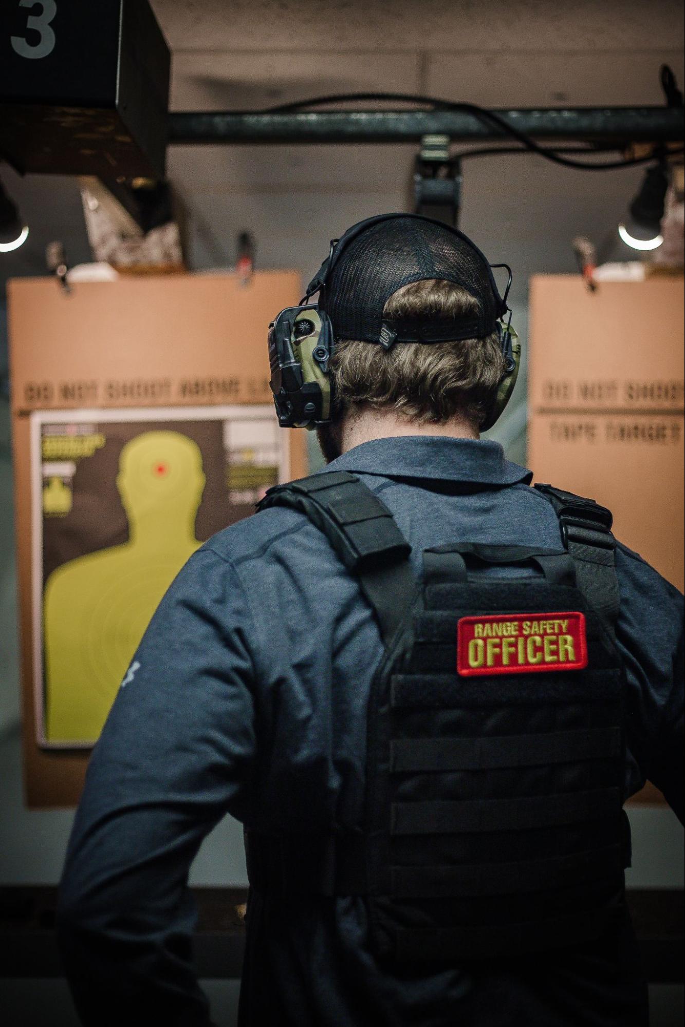 A Range Safety Officer in tactical gear faces shooting targets at an indoor range. Visible patch reads "RANGE SAFETY OFFICER."
