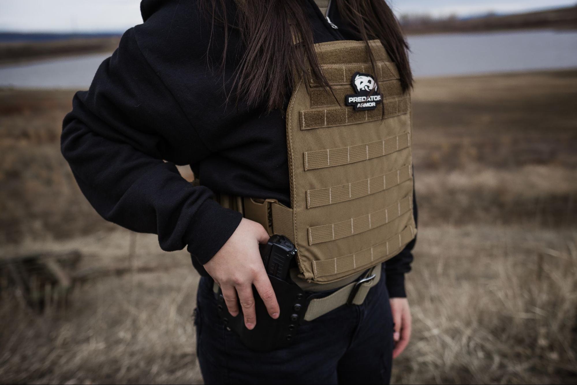 Female in a black hoodie wearing a Coyote Predator Armor plate carrier with a pistol on her battle belt. 