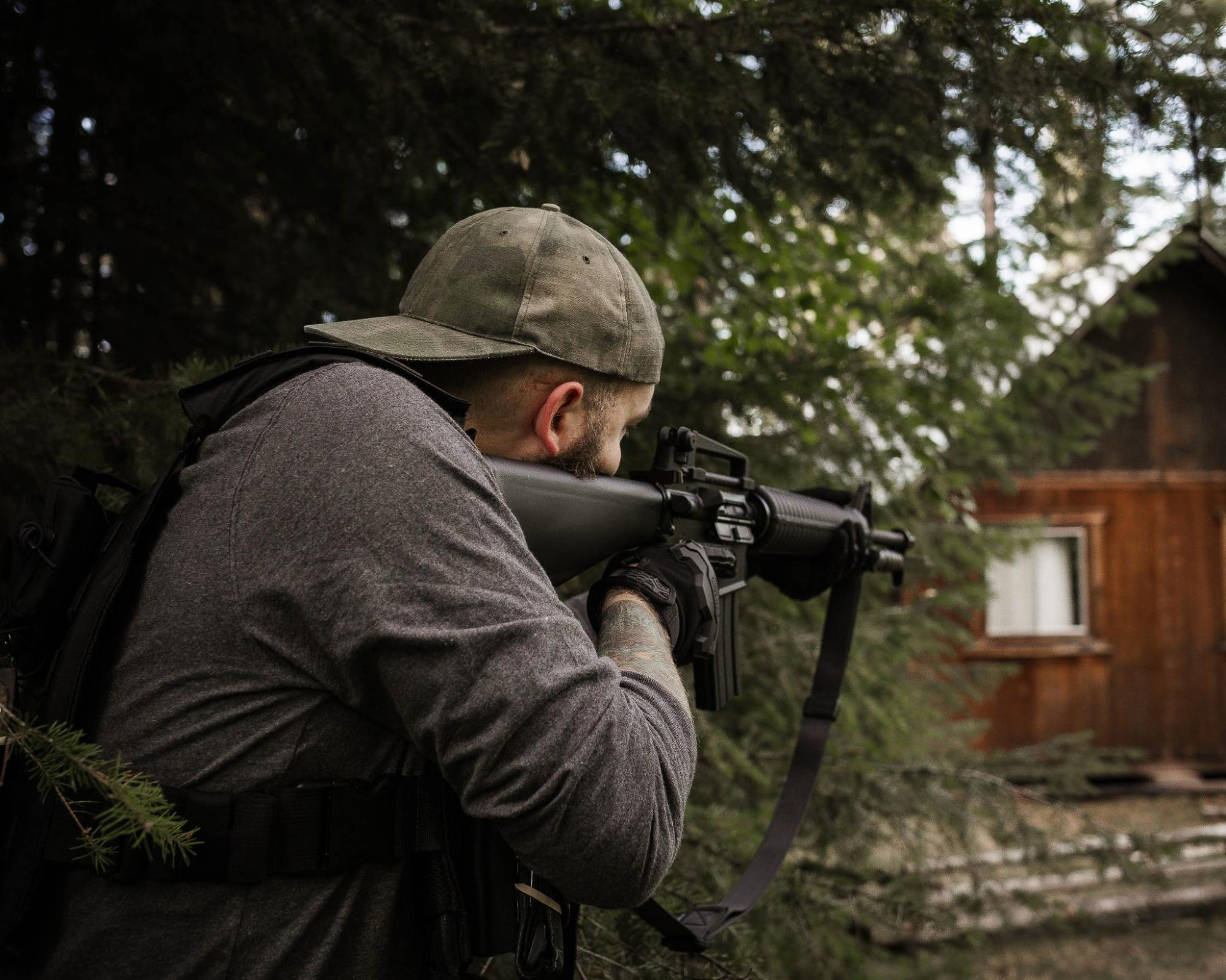 Man in cap aiming rifle, crouched among trees near wooden cabin. Wearing gray shirt, gloves, and backpack.