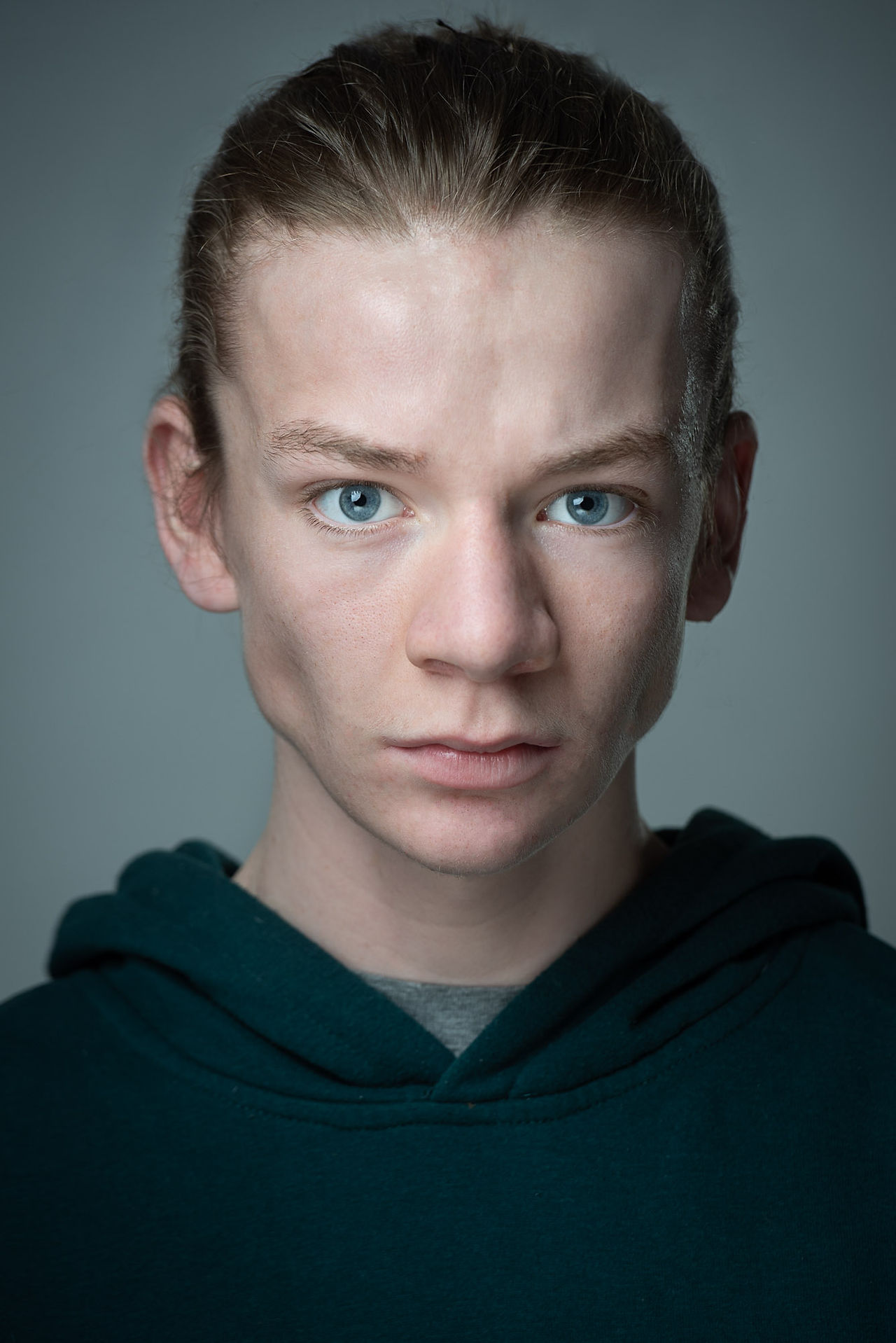 Actors Headshot, intense close up of a young performer with long brown hair tight up behind and blue eyes on light grey dark background