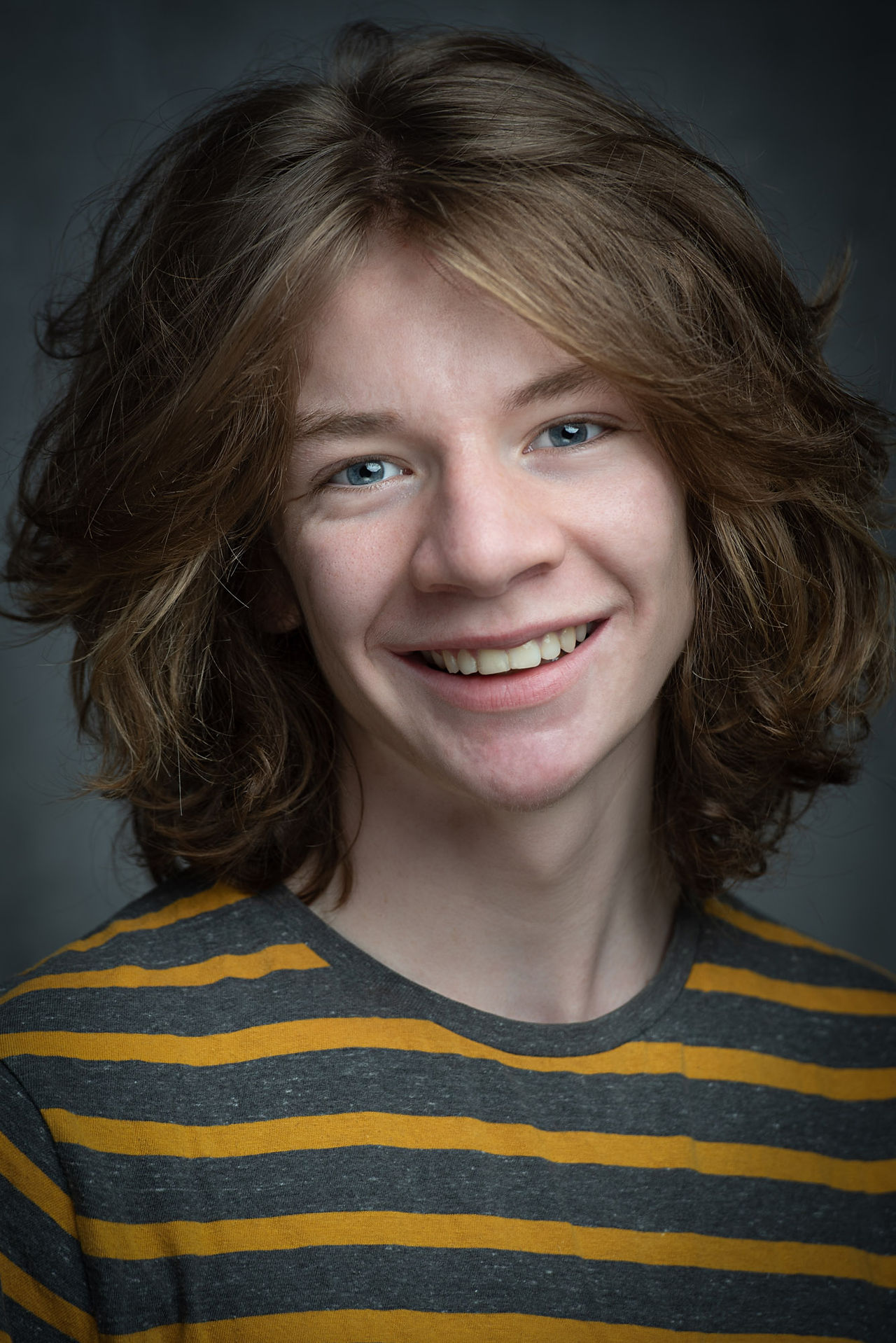 Actors Headshot, cheerful close up of a young performer with thick long brown hair and blue eyes on light grey background