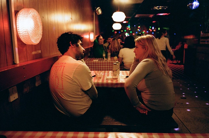 A man and a woman sitting in a bar drinking cans of beer, with other people in the background. The tables are covered with a red and white check tablecloth and clourful lights behind. The walls are 1970's style wood panelling.