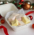 קופסת קרטון לבנה פתוחה עם קוביות מרשמלו בעבודת יד בצבעי צהוב, ורוד ולבן, המונחות על נייר אפייה מחוספס. סביב הקופסה קישוטי חג