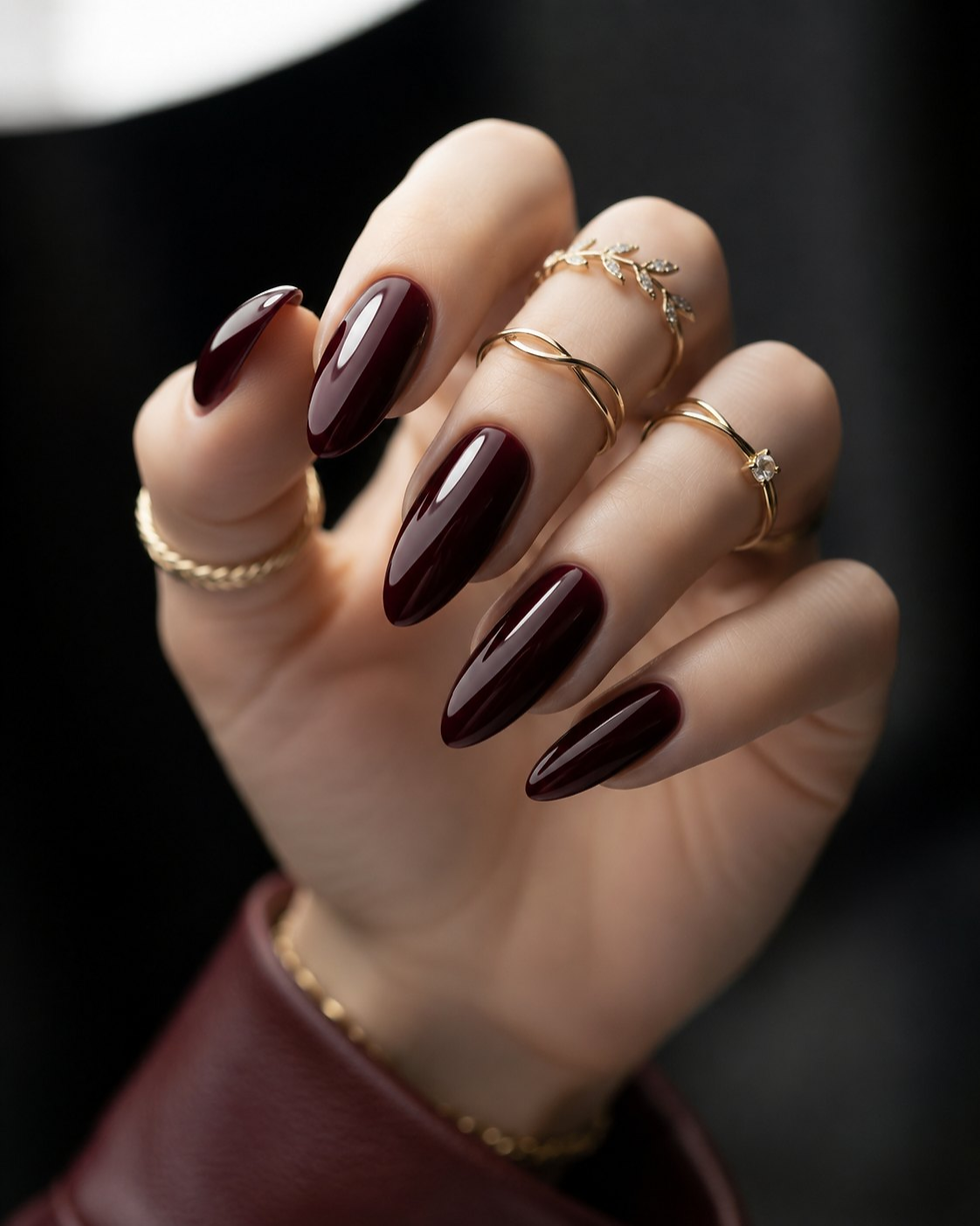 Glossy dark cherry almond nails on a hand wearing delicate gold rings