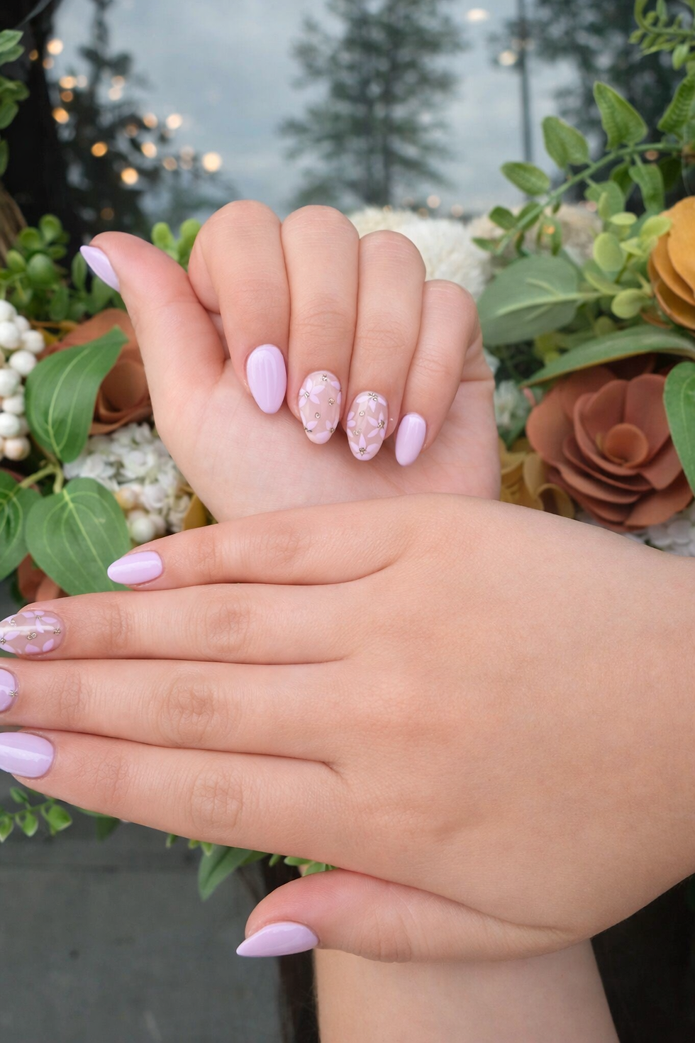 Hands with lilac and floral nail art, set against a backdrop of green and brown foliage, creating a fresh and elegant look.