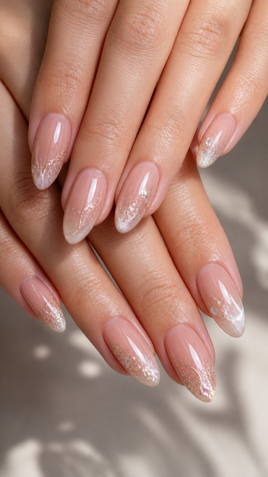 Close-up of hands with long, tapered nails featuring a pink and glitter gradient manicure. Soft lighting creates a serene mood.