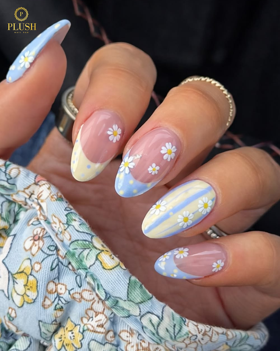 Close-up of nails painted with pastel blue, yellow, and white daisy designs. The hand holds fabric with a floral pattern.