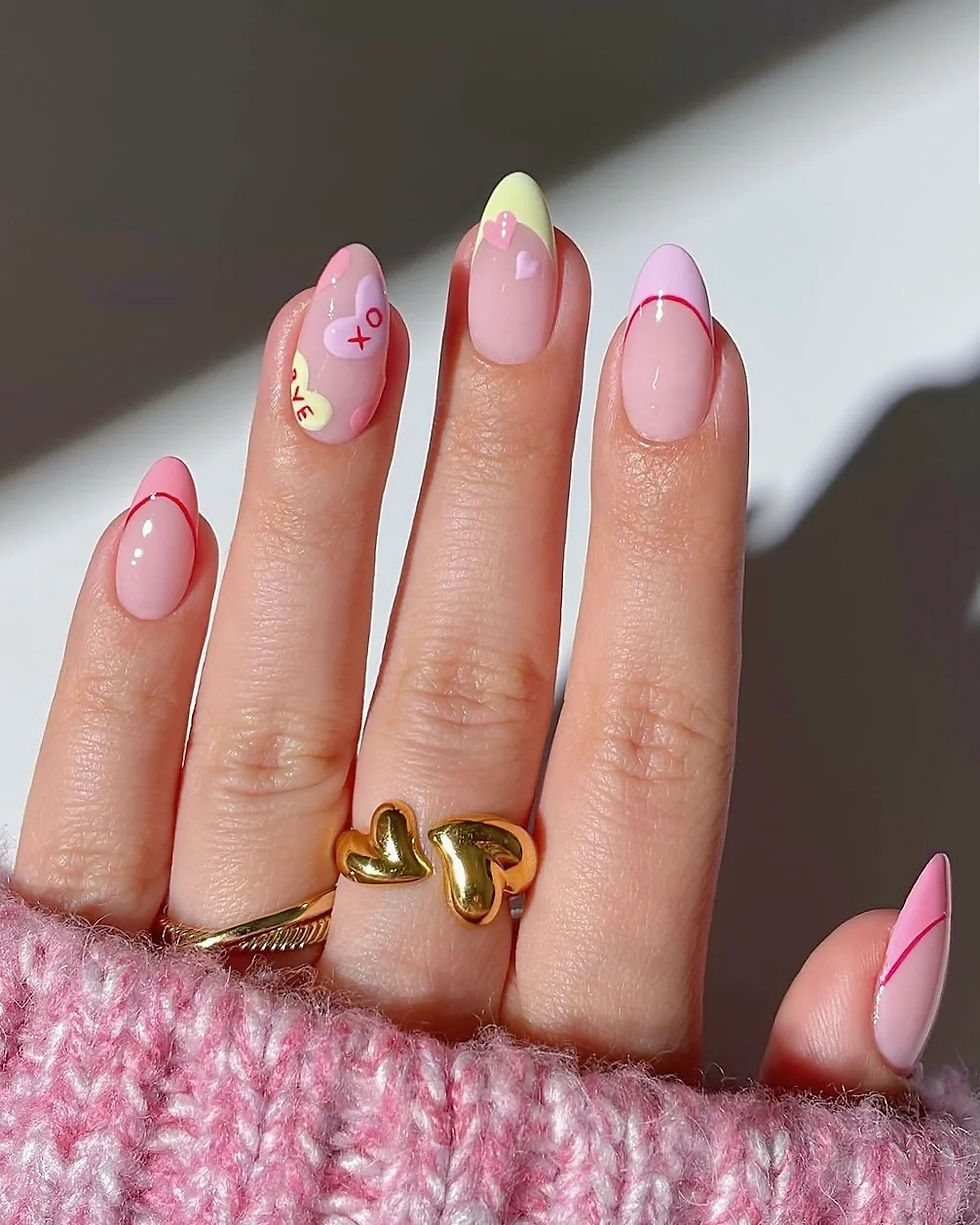 Hand with pink and yellow nail art featuring hearts and "XO" text. Gold heart ring on finger. Knitted pink sweater in background.
