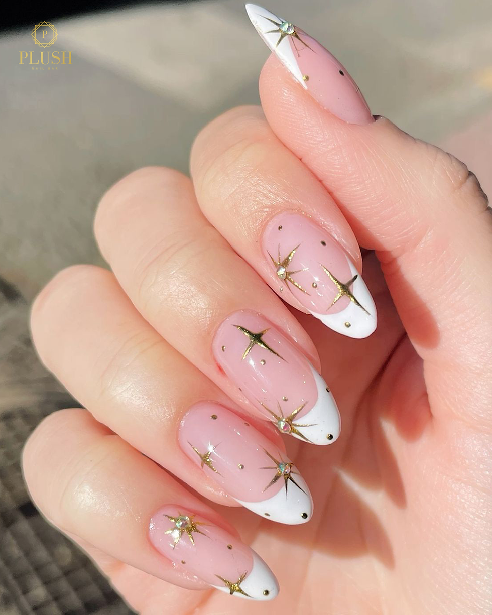 Pink nails with white tips and gold star designs, hand held up in natural light. Text "PLUSH Nail Bar" in top left corner.