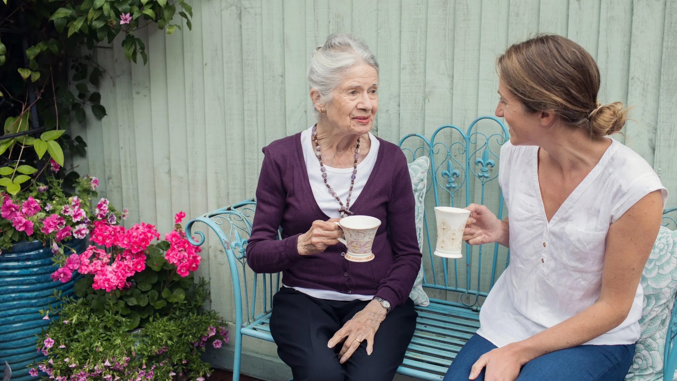 Home carer sat with elderly lady drinking in pretty garden