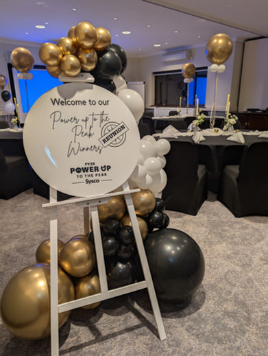 White circular branded welcome disk on white easel frame and gold, black and white balloons. 