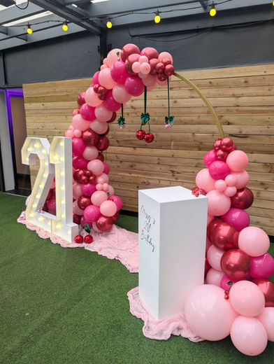 A cherry theme red and pink balloon hoop, mirror ball cherries hung from the centre, light up 21 marquee numbers and personalised white wooden cake plinth.