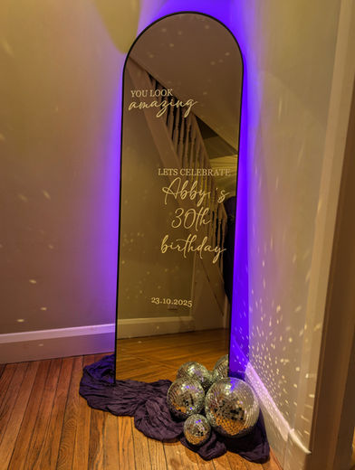Curved mirror with white personalised text, thin black frame, mirror balls at the base and a purple uplighter behind.