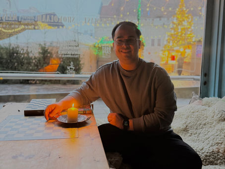 A person smiles while sitting inside, lighting a candle on a wooden table with a checkerboard. Festive lights and village scene visible outside.