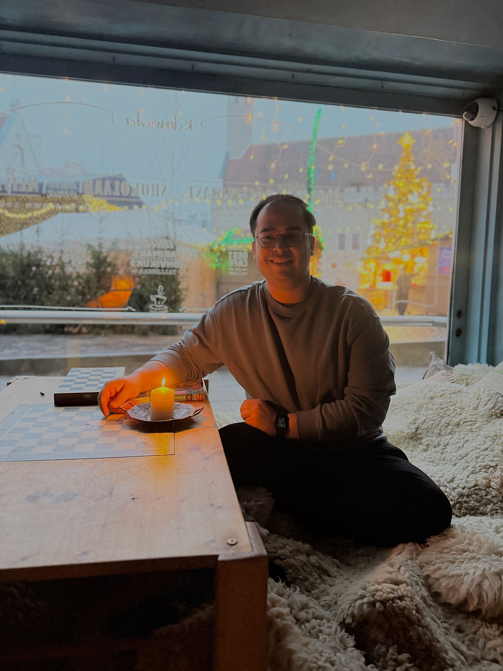 A person smiles while sitting inside, lighting a candle on a wooden table with a checkerboard. Festive lights and village scene visible outside.