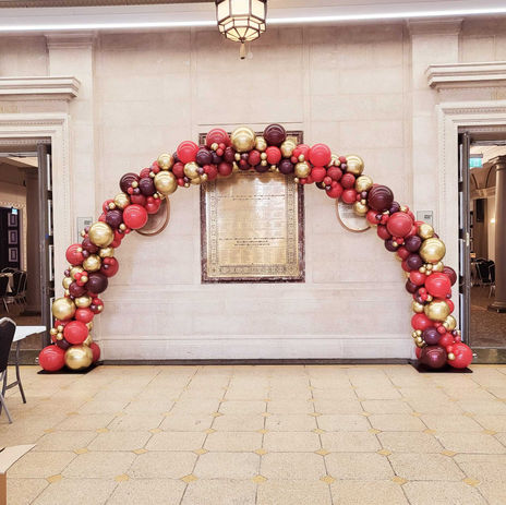 An organic balloon arch made out of red, purple and gold balloons, placed in a grand hall with a gold, engraved plaque behind it.
