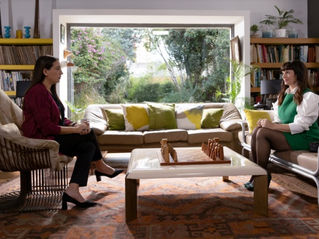 Cena da série "Envidiosa", a psicóloga sentada na poltrona ao lado esquerdo de frente para a sua paciente Victoria, mulher, sentada ao lado direito, com uma janela no meio das duas mostrando um jardim ao fundo. Um consultório com livros, almofadas e prateleiras amarelas.