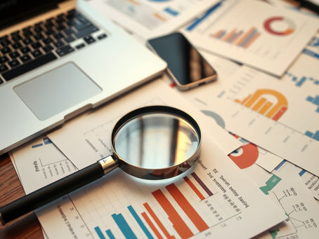 An office desk covered with papers showing graphs, charts and tables. A laptop is open on the desk and a cellphone sits next to it. A large magnifying glass sits on the pile of papers.