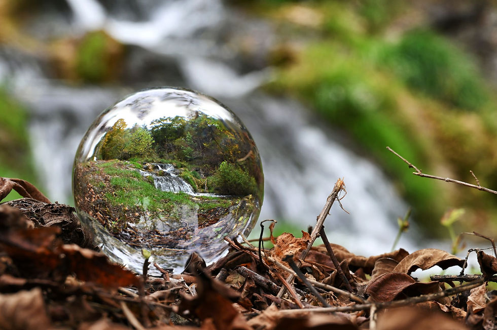 A woodland scene in which a crystal ball sits amongst some leaves and twigs. The view through the crystal ball is a crisply focused waterfall that is out of focus in the distance of the overall scene.