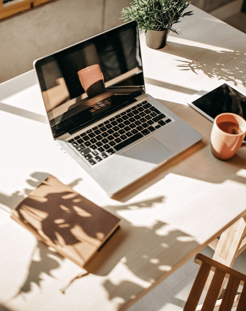 Imagem com notebook, agenda, um celular a direita do aparelho e uma caneca refletida na tela