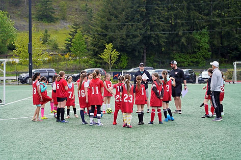 A photo of middle-school aged girls with numbered pinnies at tryouts, standing around listening to coaches.