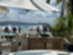 A photo of a beautiful tropical resort. The vantage point is a casual dining table overlooking other tables with umbrellas. Beyond is a tropical blue ocean.