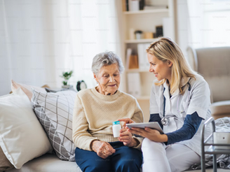 A nurse shows a tablet to an elderly woman holding a pill bottle. They're seated on a couch in a bright, cozy room, smiling at each other.