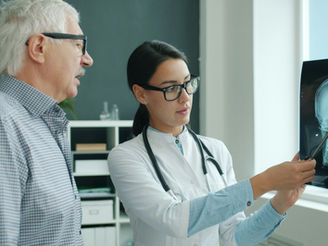 Doctor with stethoscope shows brain X-ray to older man, pointing with pen. Office with shelves in background, conveying a professional mood.