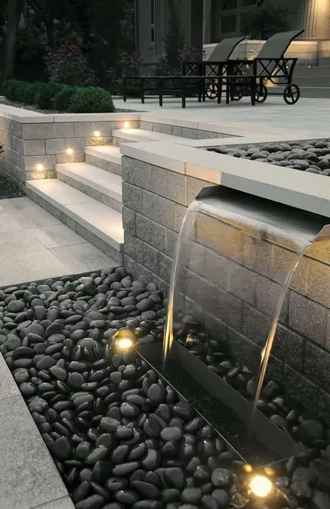 Modern patio with illuminated steps and a water feature flowing over stones. Lit areas create a calm ambiance with seating in the background.