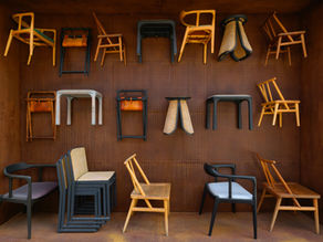 A straight on view of a deeply textured warm rust colored pegboard wall creatively displaying a curated variety of artisan chairs and stools. The top two rows appear to float against the rich brown background featuring smooth curved light oak frames matte black painted seats woven rattan stools and folded leather camp frames. Resting on the matching floor below is a neat arrangement of seating including a stacked set of black cane dining chairs alongside elegant wooden armchairs with soft grey fabric cushions.