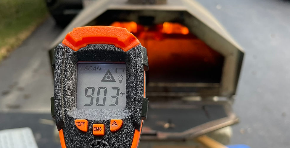 Infrared thermometer shows 903°F in front of a lit pizza oven. The thermometer is black and orange, set outdoors.