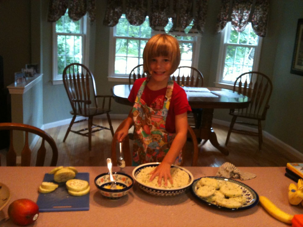 Emma in colorful apron smiles while preparing food in a kitchen, with sliced eggplant on counter. Sunlit windows and wooden chairs in background.