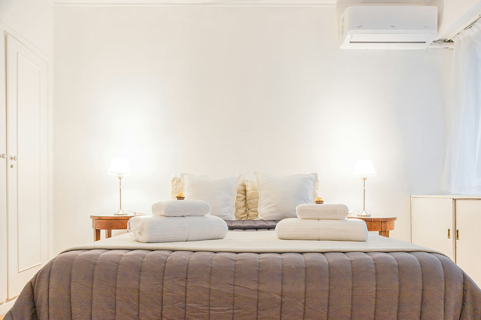 Serene hotel bedroom with neutral color palette, showing a bed dressed in a taupe channel-quilted comforter with white pillows and rolled towels arranged on top, flanked by wooden nightstands with modern white table lamps, white walls, and a mini-split air conditioner mounted above