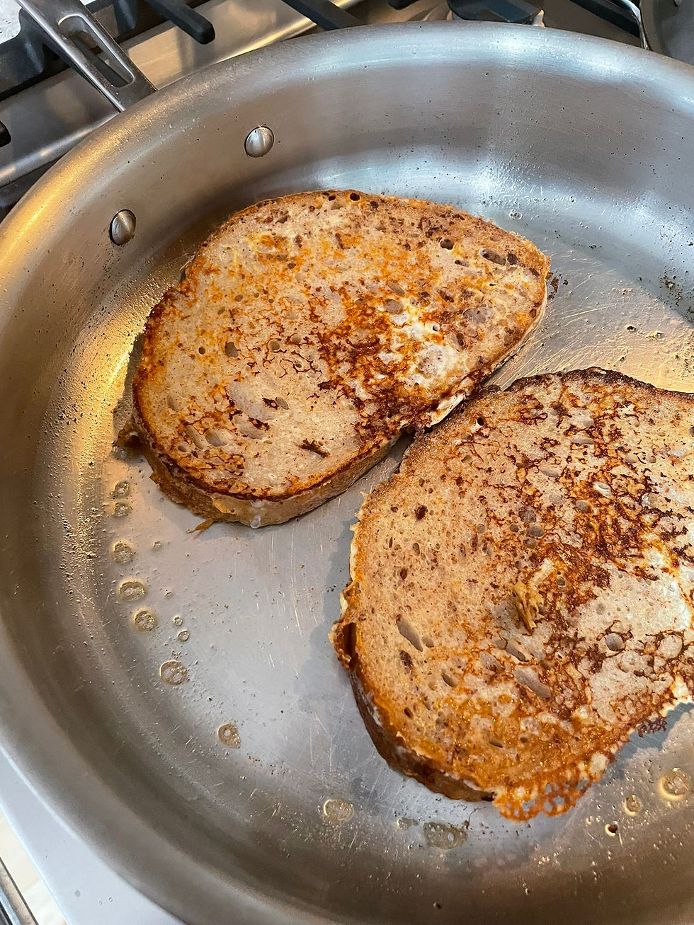 Two slices of golden brown toasted bread with seeds frying in a silver pan on a stovetop, with bubbles of oil visible.