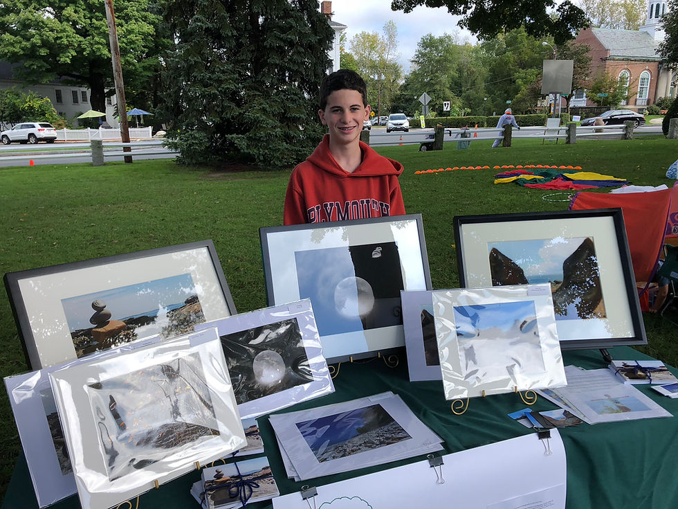 A young artist with a bright smile stands proudly behind an outdoor wooden table draped with a rich green tablecloth. Framed nature photography prints in polished black and silver borders sit neatly on display under the warm open air. The crisp scent of fresh grass mingles with the faint earthy aroma of damp soil and sun-warmed pine needles. Sunlight gently bounces off the protective plastic covers of the photographs, casting a soft, shimmering glow across the entire booth.