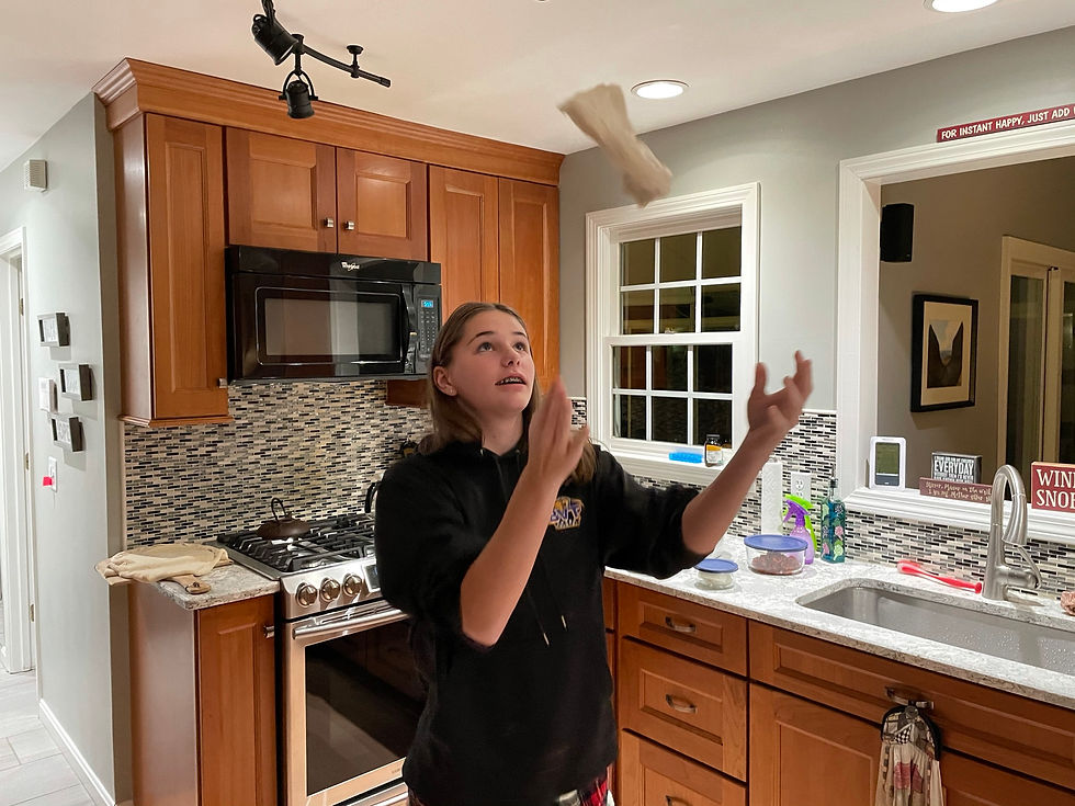 Emma in a black shirt stands in a modern kitchen with her hands raised in the air, appearing to be juggling or catching something. The kitchen features warm wood cabinets, granite countertops, a mosaic tile backsplash, stainless steel appliances including a microwave and gas range, and a window with white trim. Various items are visible on the counter including bowls and kitchen accessories.
