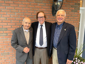 A full medium shot of three lifelong friends standing shoulder to shoulder outdoors in front of a red brick wall. The man on the left has a bald head and wears a grey patterned blazer over a cream collared shirt. The man in the center wears black rimmed glasses, a white dress shirt, a dark patterned tie, and grey trousers. The man on the right has short white hair and wears a dark blue blazer over a blue and white plaid button down shirt with khaki pants.