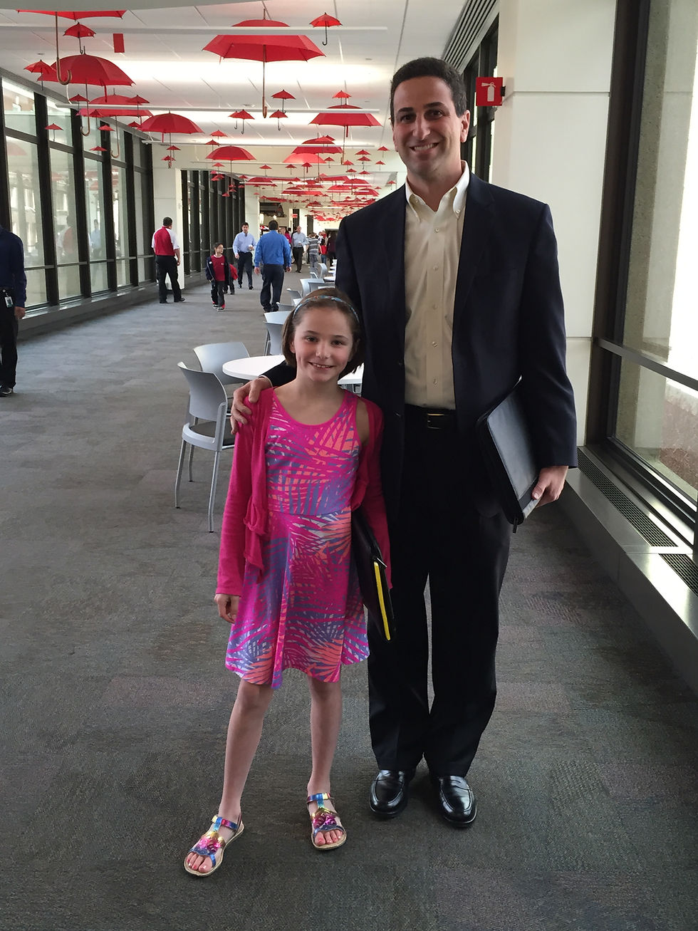 A wide-angle photograph of a smiling man, Steve Fiore,
in a navy blue suit and dress shoes standing with his arm around a young girl, Emma, in a bright pink and purple patterned dress, in a long hallway with floor-to-ceiling windows and a unique ceiling installation of suspended red umbrellas, as people walk in the background.