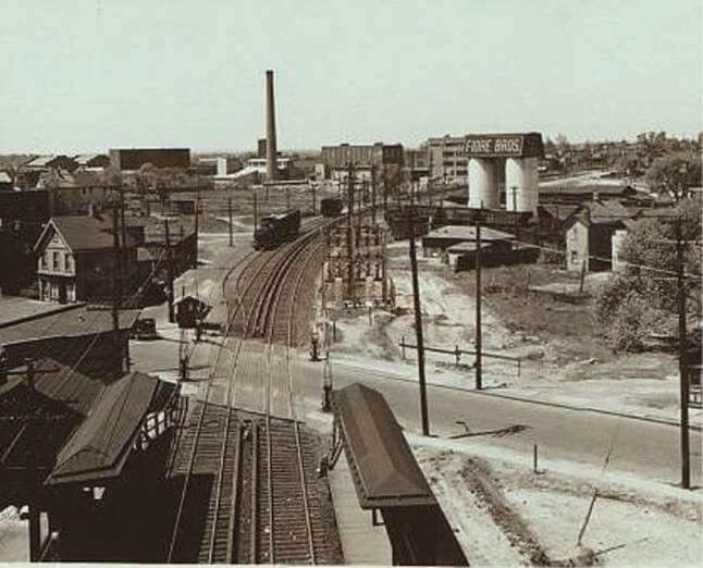 A sepia-toned historical photograph of the Fiore Bros. industrial site, featuring towering white silos and steel railroad tracks cutting through a gritty, smoke-filled cityscape in Staten Island, NY.
