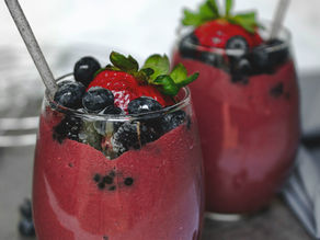 Two glasses filled with vibrant pink-purple berry smoothies topped with fresh blueberries and strawberries with green leaves, served with metal straws on a gray surface