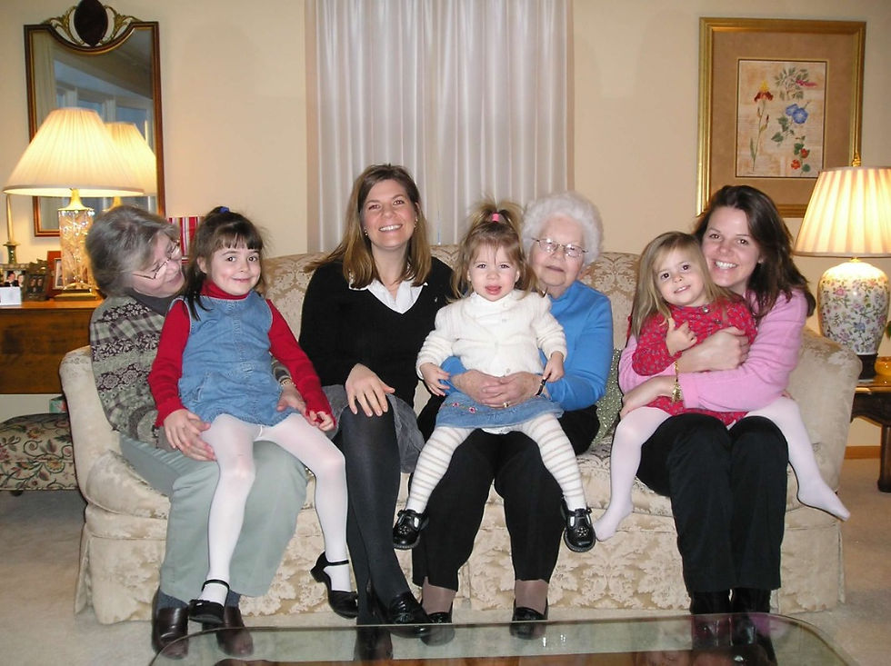 This indoor photograph captures four generations of females arranged tightly together on a classic beige couch with a subtle floral damask pattern. On the far left, an older woman with short grey hair and glasses leans inward, smiling warmly at a young girl in a blue denim jumper, red turtleneck, and white tights sitting on her lap. Next to them sits a woman in a black v neck sweater over a white collared shirt and dark tights, smiling directly at the camera. In the center, an elderly matriarch with short, voluminous white hair, glasses, and a bright blue long sleeve top holds a toddler on her lap. The toddler has her hair in a high ponytail, wearing a white knit cardigan, a denim skirt, and striped leggings. To the right, another young girl in a red floral dress and white tights is held tightly by a woman in a pink sweater and black pants, both flashing bright smiles. The room is illuminated by two large traditional table lamps with pleated fabric shades emitting a soft, golden glow. A framed floral painting hangs on the wall to the right, and a large mirror on the left reflects the warm room. The image conveys a sense of soft textures, from the plush fabric of the sofa and the crisp cotton collars to the cozy knit of the white sweater and the smooth feel of children's tights. The atmosphere feels physically warm, crowded, and affectionate.