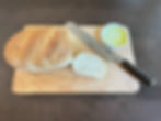 Loaf of bread and sliced piece on wooden board with knife and small bowl of yellow butter. Dark table surface creates a cozy mood.
