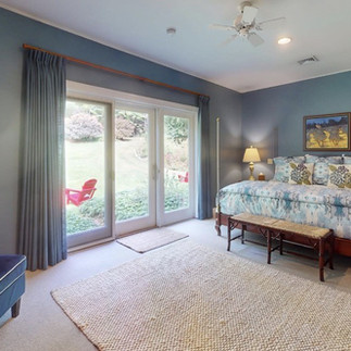 A tranquil bedroom bathed in bright natural light streaming through large sliding glass doors. The doors look out onto a vibrant green lawn where a bright red wooden chair sits. Inside the room features calming blue walls and a large bed dressed in soft blue and white patterned linens. A deeply textured woven rug covers the floor adding a warm tactile element to the space.