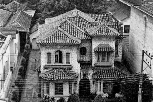 Foto em preto e branco. A imagem apresenta uma vista superior da Residência Ekman. Possui uma marca d’água no canto inferior direito com os escritos “www.Maria do Resguardo.com.br”. São visíveis, além da casa, o restante do lote com o acesso à garagem nos fundos, à esquerda, e o quintal. Em primeiro plano, há pessoas transitando no passeio em frente à casa, algumas se apoiam no muro frontal composto por três portões, o da extremidade esquerda, é baixo e largo.O acesso à garagem nos fundos do lote é profundo. O portão do centro é baixo e possui uma cobertura em dois caimentos. O terceiro, na extremidade direita do muro, também é baixo e possui uma estrutura arqueada acima. Atrás do muro, em frente à casa, há uma pequena vegetação seguida da fachada frontal da residência, toda ornamentada e composta por dois volumes principais ressaltados e outros dois volumes recuados, os intercalando. No primeiro pavimento, a entrada da casa é composta por um vão arqueado. As janelas e a varanda possuem coberturas de telha. No segundo pavimento uma das janelas também possui uma pequena cobertura. A fachada possui uma faixa com ornamentos parecidos com arabescos próxima ao telhado e marcas decorativas por toda a sua extensão. A cobertura possui diferentes volumes e caimentos. O quintal, atrás da residência, possui uma vegetação alta. Ainda na imagem é visível parte das edificações vizinhas nas laterais e, ao fundo, outras construções.